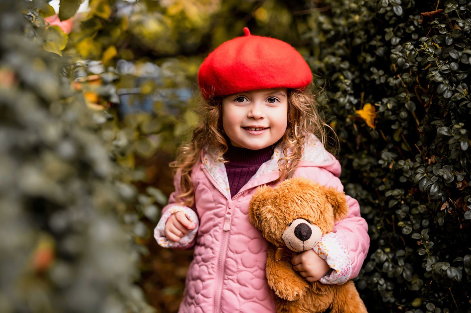 Child in pink coat and red beret holds teddy bear against garden foliage.
