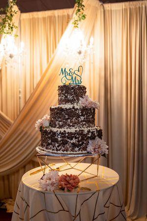 Elegant three-tier chocolate wedding cake with white flowers displayed against draped fabric backdrop.