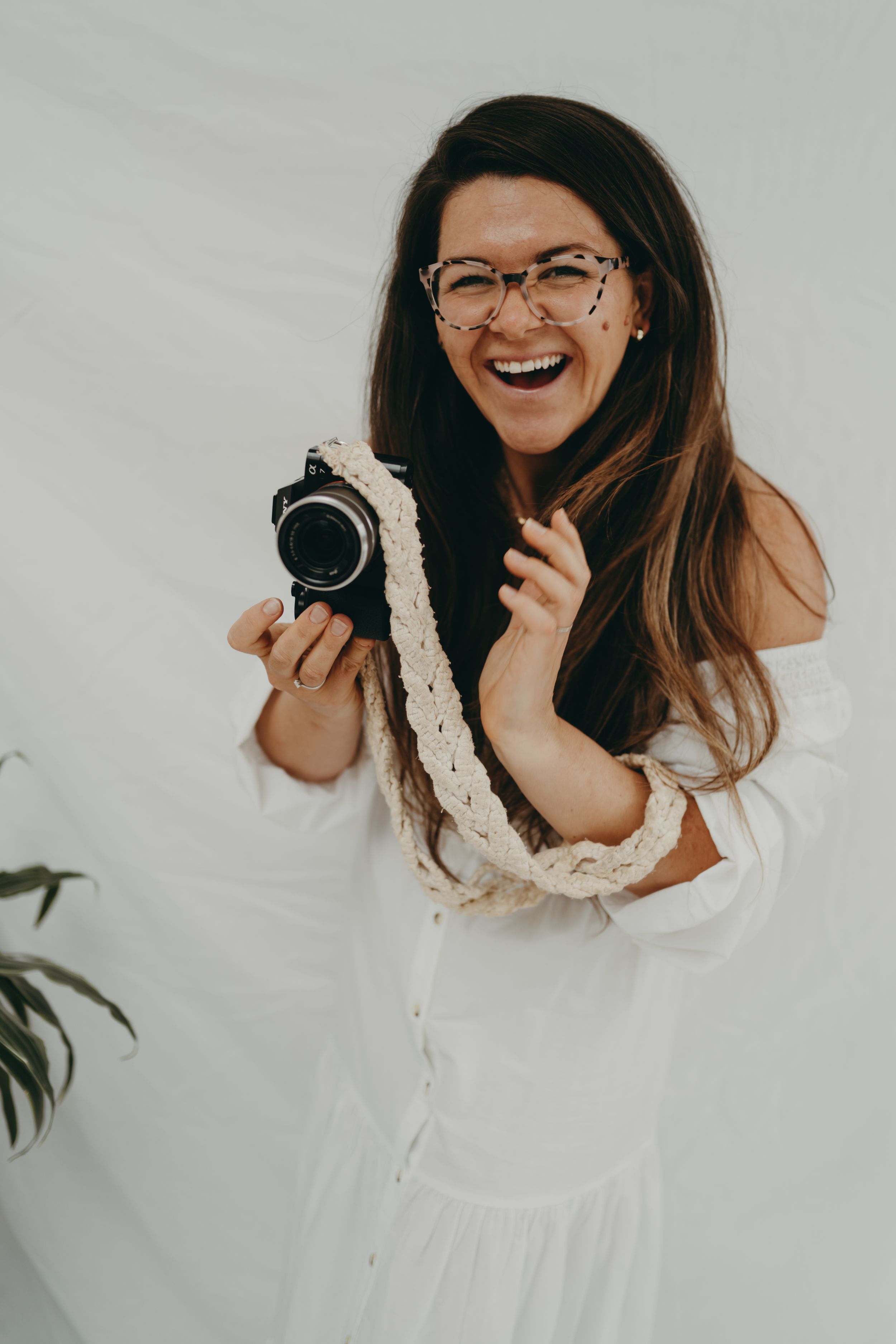 Joyful photographer in white dress holding vintage camera against light background.