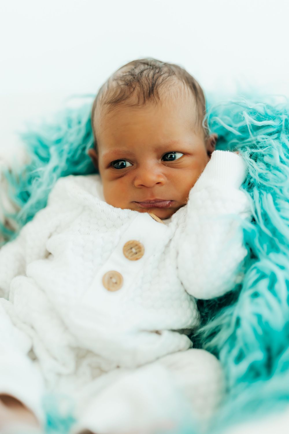 A baby wearing a white knit outfit with wooden buttons lies on a turquoise textured blanket or wrap.