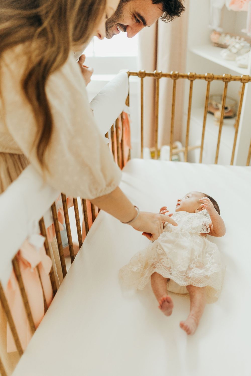 Mother in white dress bottle feeding newborn baby in wooden crib in soft natural light.