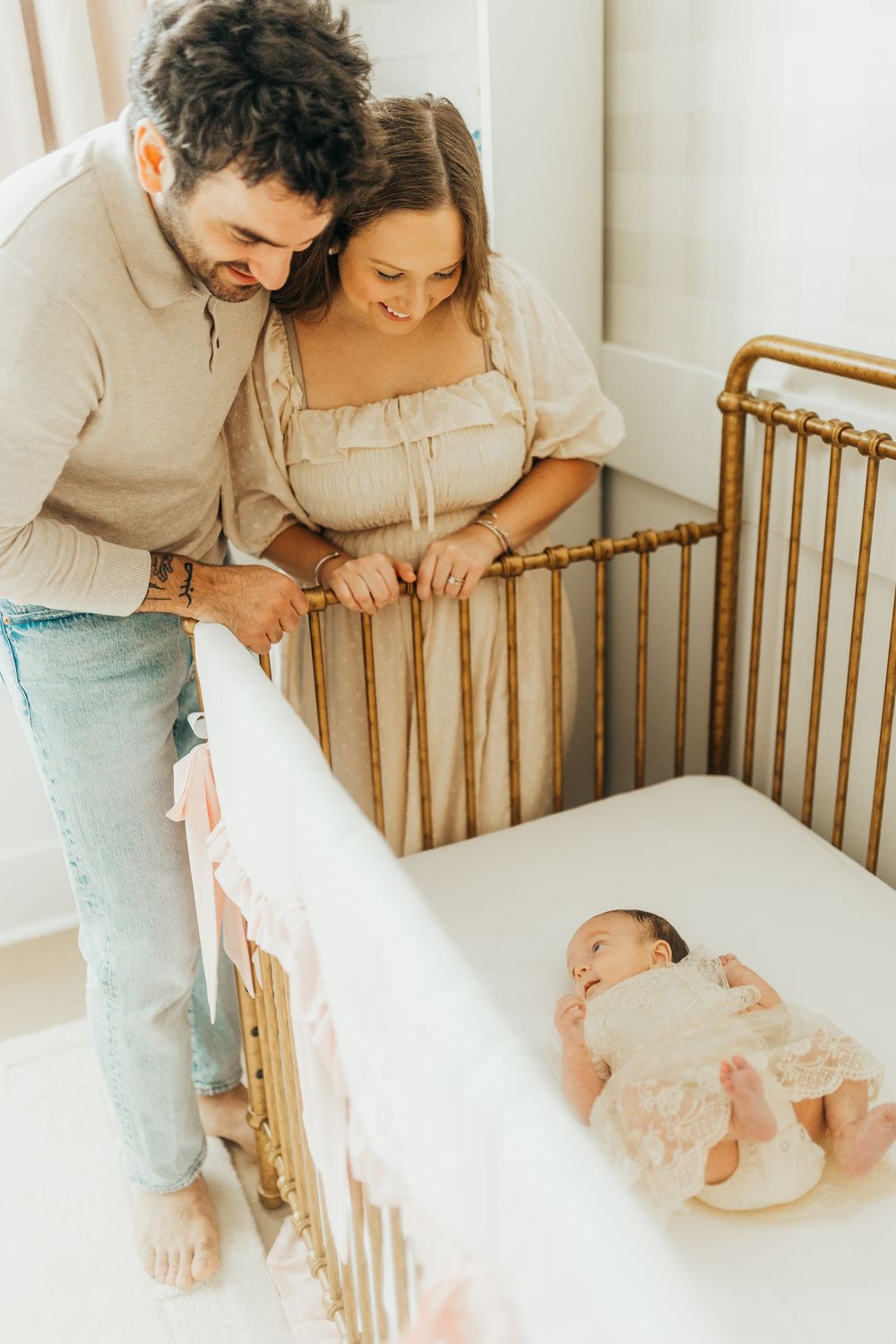 Parents admiring their newborn baby lying in a wooden crib wearing white clothing.