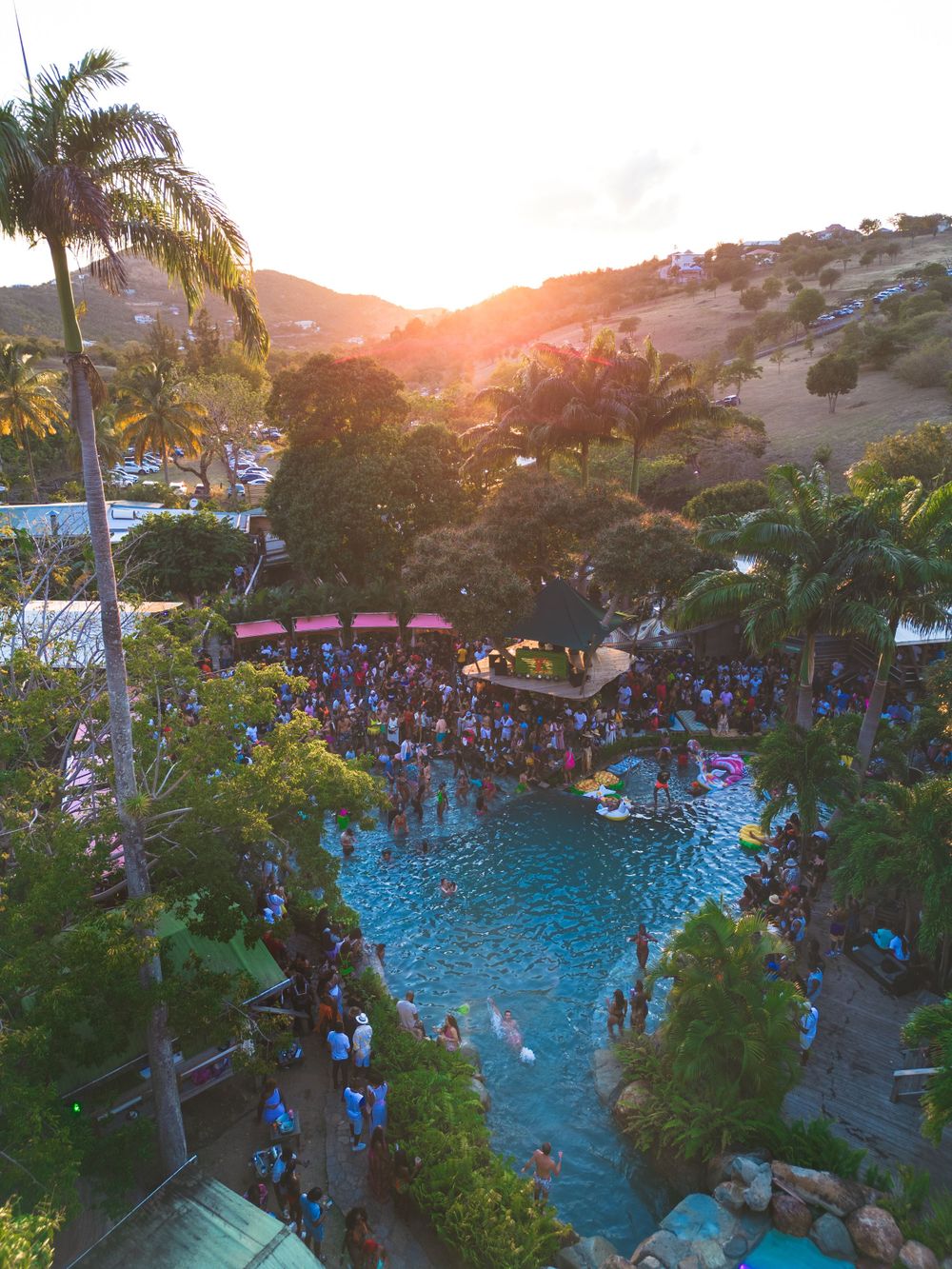 Aerial view of a packed pool party at sunset surrounded by palm trees and tropical landscaping.