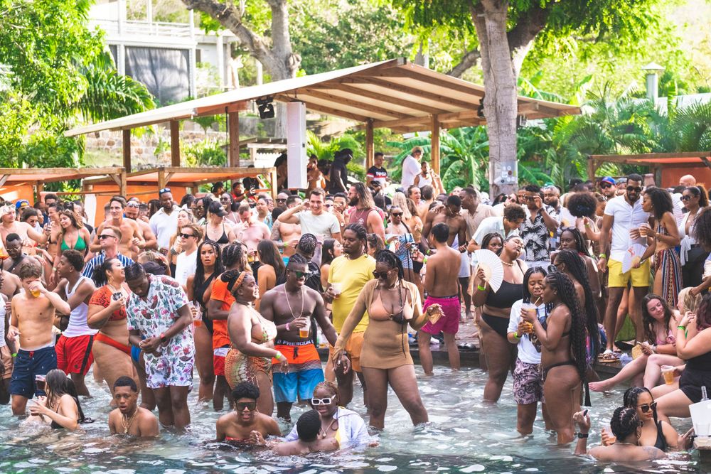 Large crowd of people enjoying a pool party at an outdoor venue with tropical landscaping and a cabana in the background.