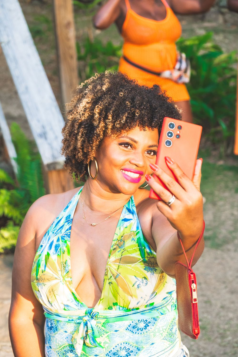 Someone takes a selfie outdoors wearing a colorful tropical print halter dress while holding a coral-colored smartphone.