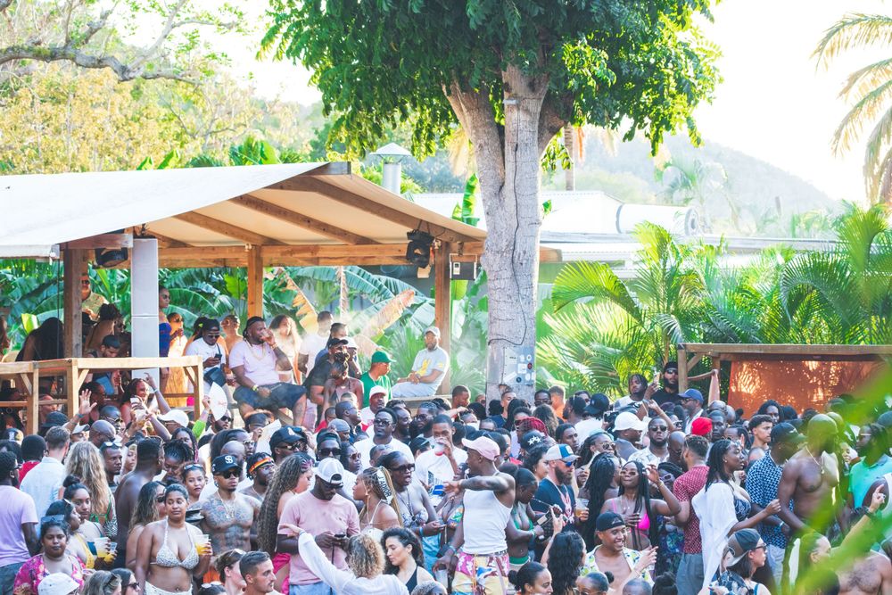 Large crowd of people gathered under covered outdoor venue area surrounded by tropical plants and palm trees in daylight.