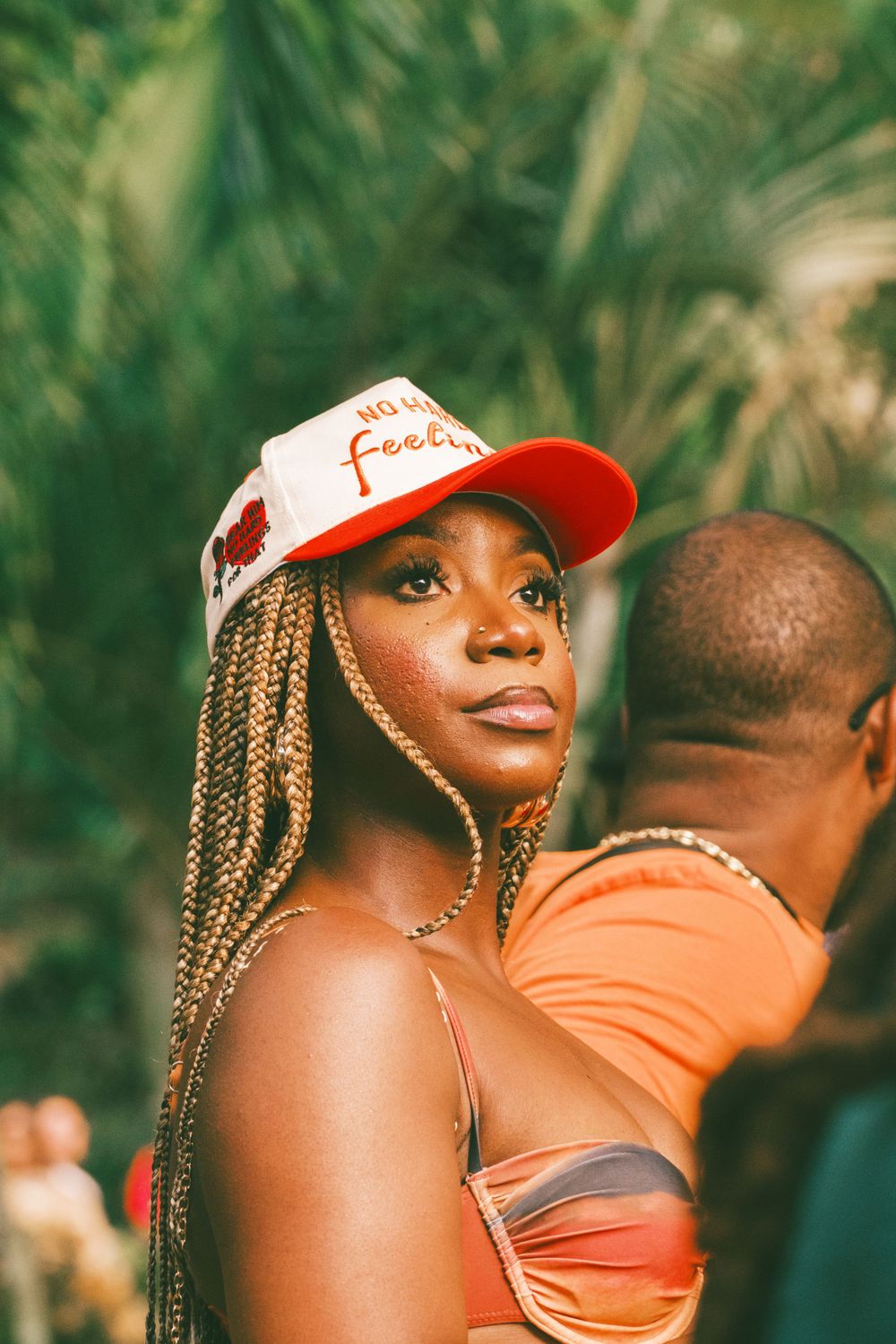 Person in red cap and braided hairstyle poses in front of tropical foliage wearing orange attire.