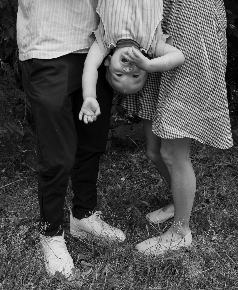 Black and white photo of a child being held upside down between two adults standing on grass.