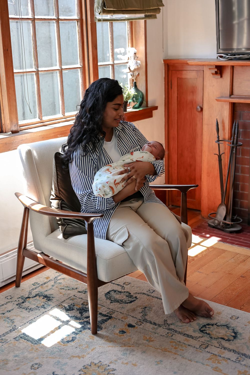 A woman sits in a mid-century modern chair holding a newborn baby in a sunlit living room with hardwood floors.