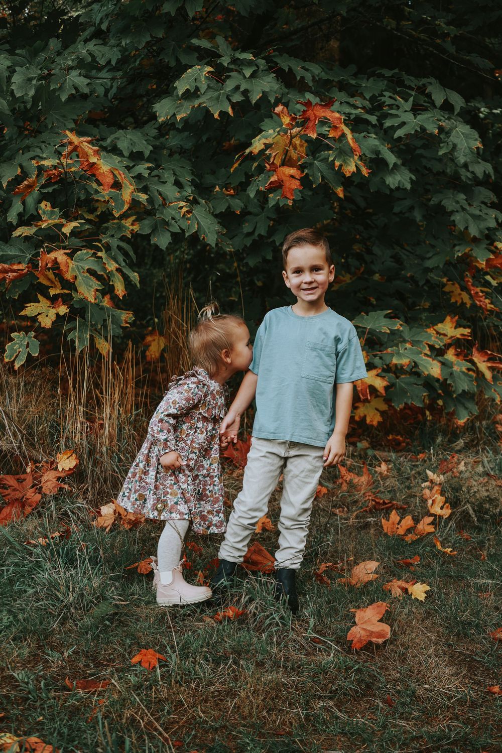 Two children stand together among fall foliage and maple trees in an outdoor autumn setting.