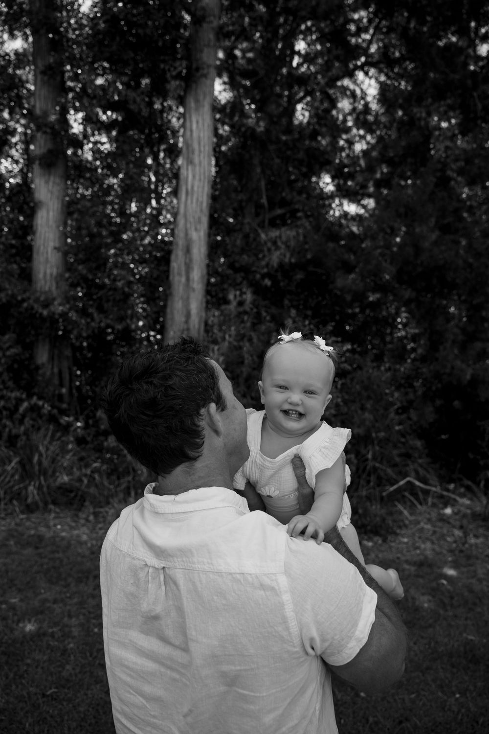 Black and white photo of a father lifting his smiling baby girl outdoors near trees.