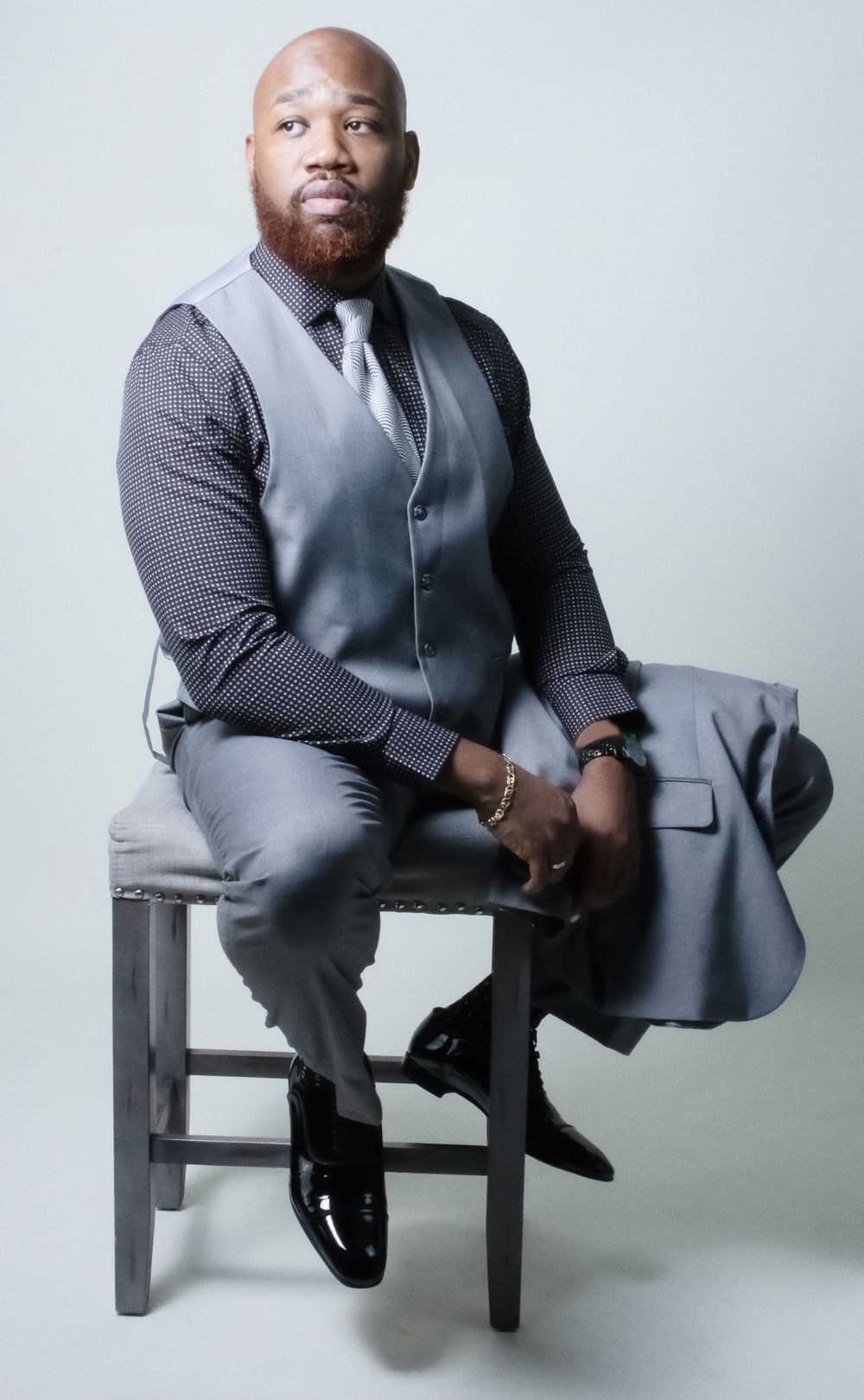 Professional studio portrait of person in grey suit vest seated on wooden stool.