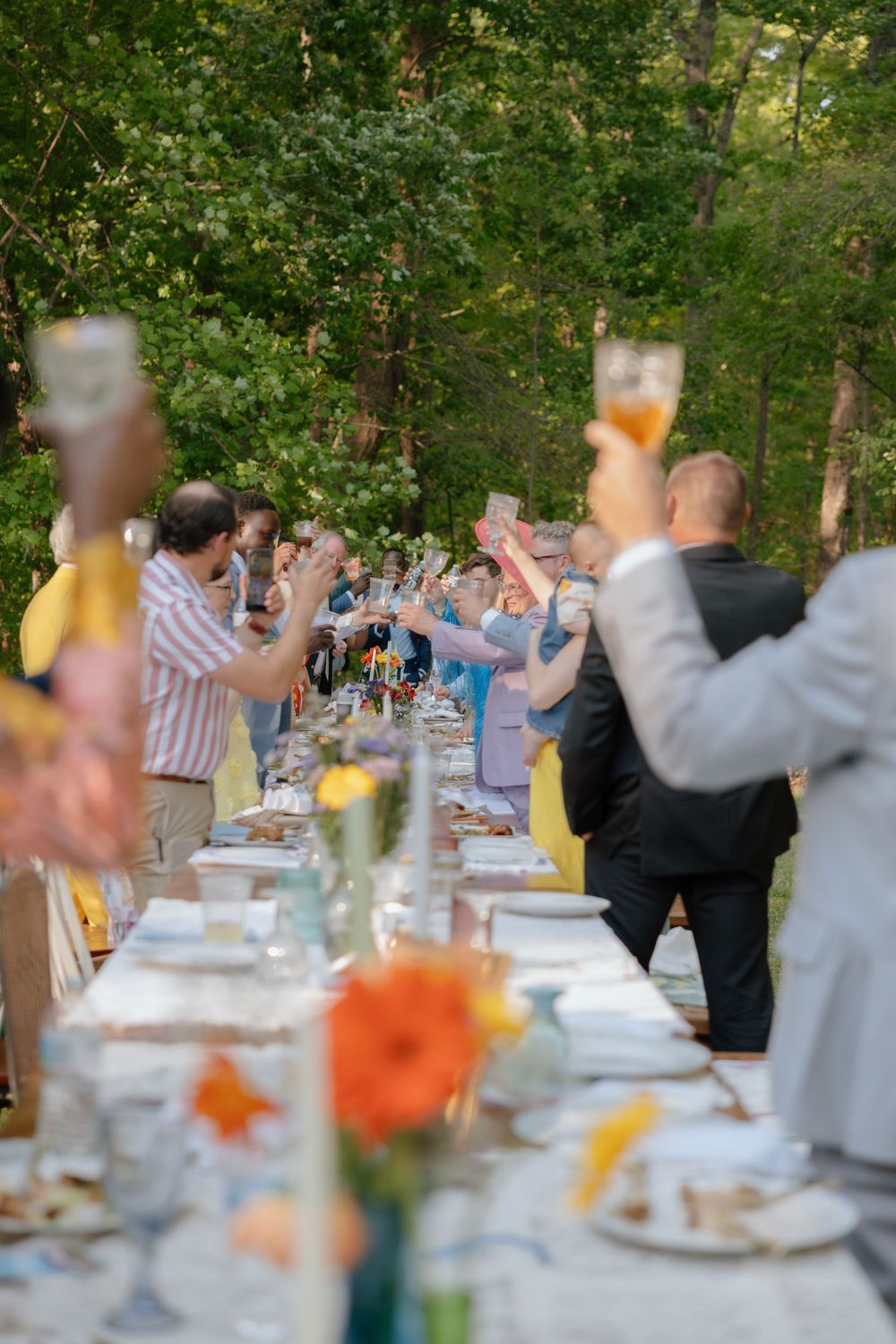 Wedding guests raise their glasses in a toast at an outdoor reception with tables decorated with orange flowers.