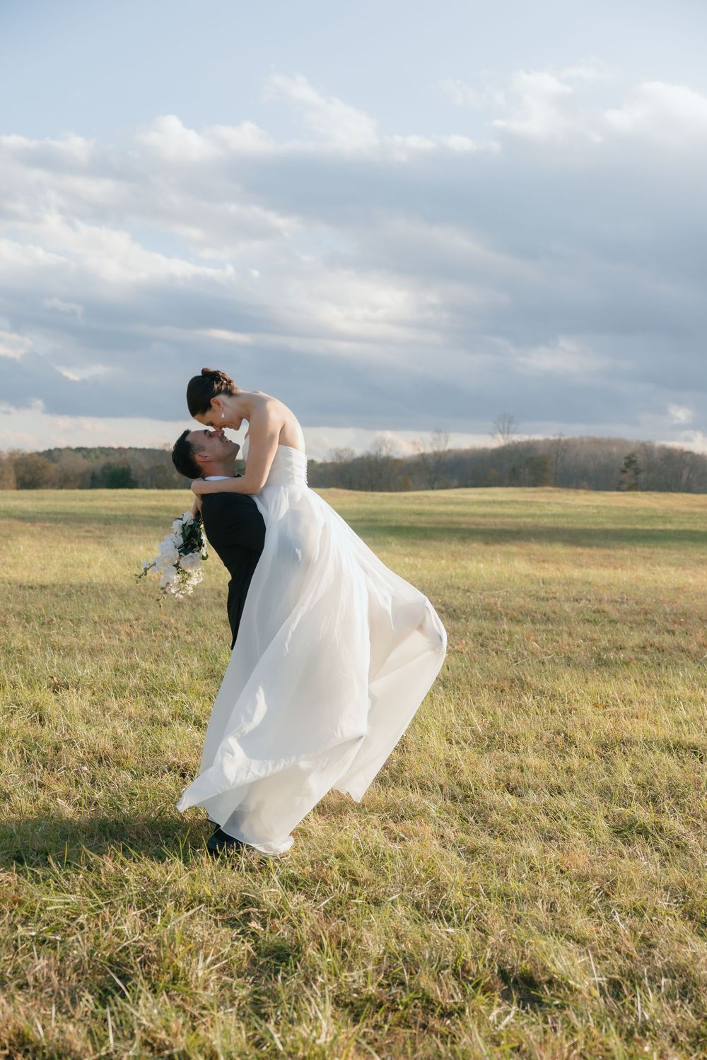A couple shares a romantic embrace in a flowing white wedding dress against a scenic countryside meadow at sunset.