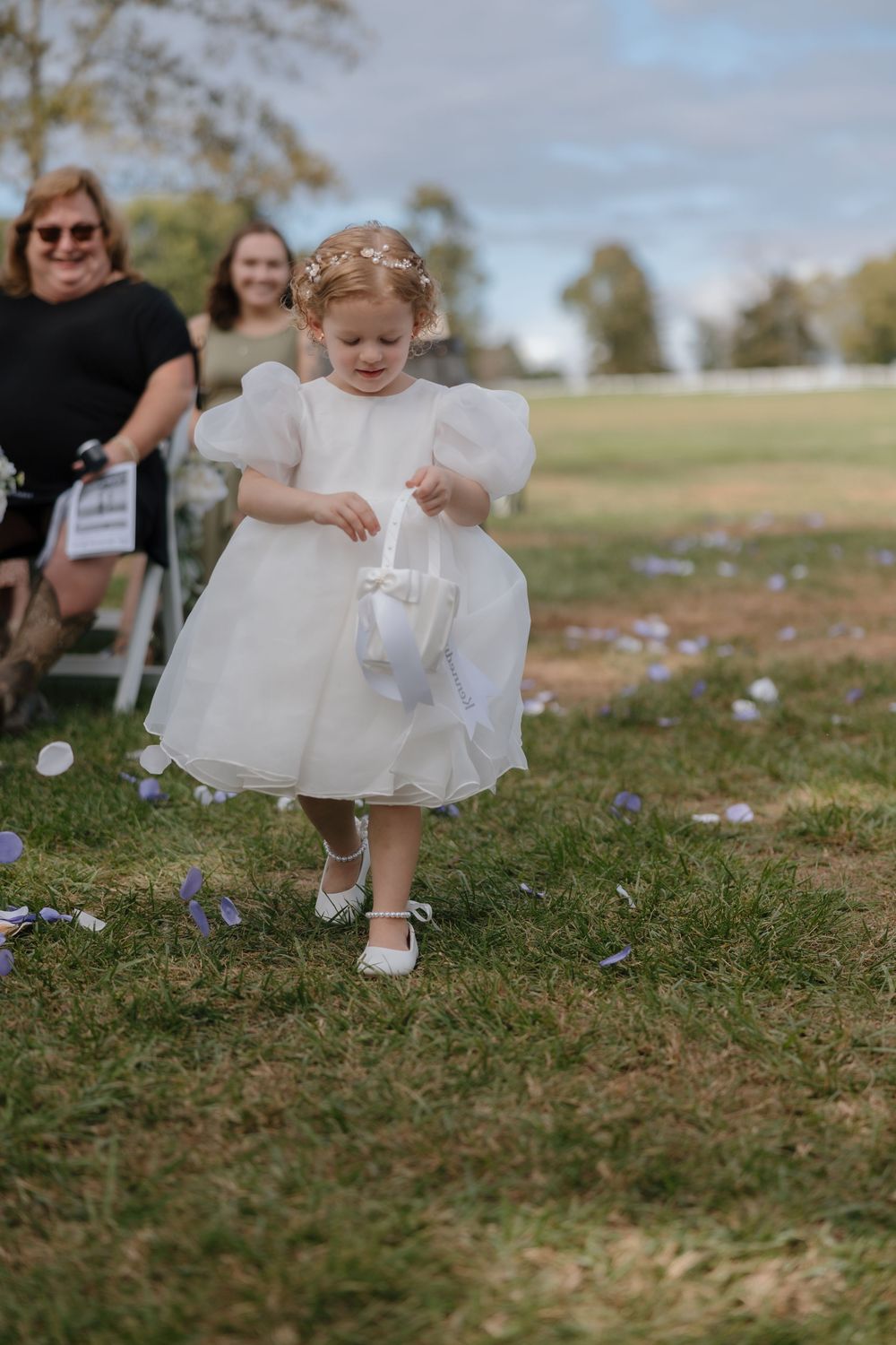 A young child in a white dress sits on grassy ground while playing with their dress hem during an outdoor event.