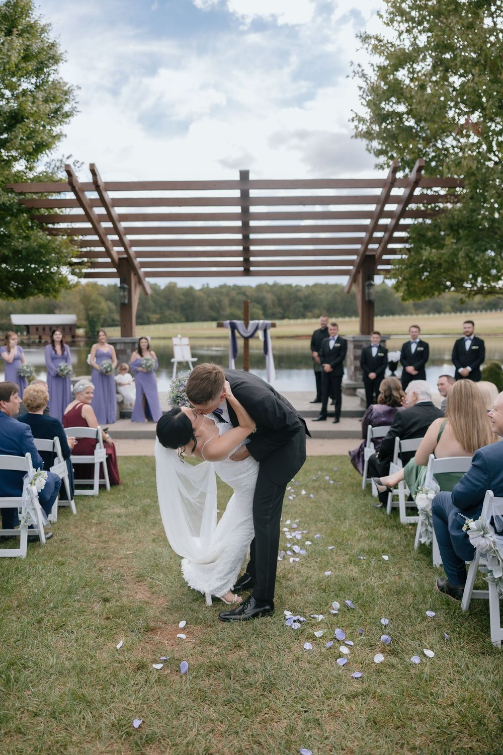 A wedding ceremony takes place outdoors under a wooden pergola with guests seated in white chairs on the grass.