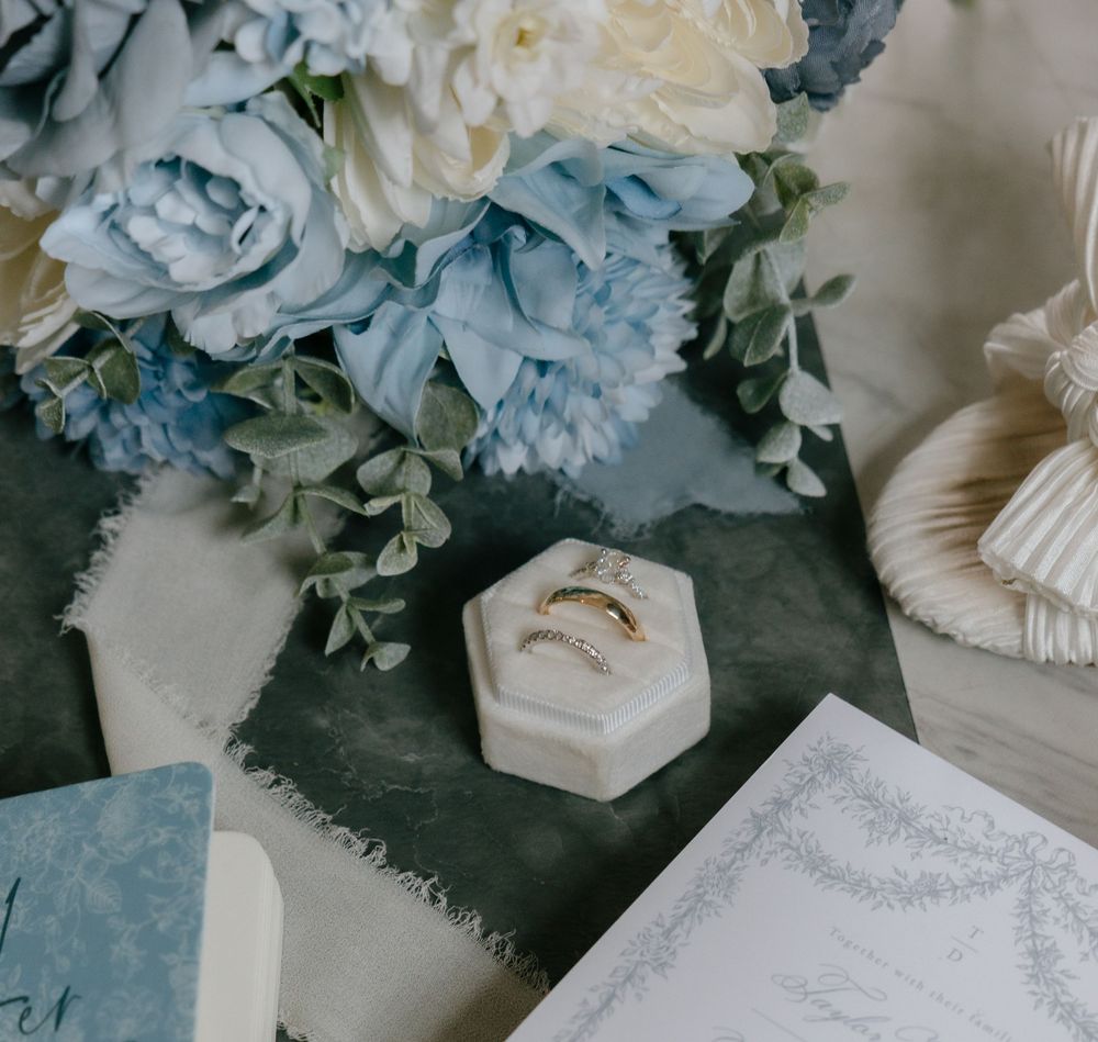 Wedding details featuring light blue floral arrangements and a white ring box on a textured gray surface.