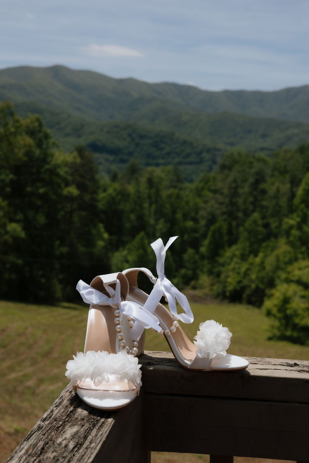 Elegant white bridal sandals with floral details sit on wooden railing against scenic mountain landscape backdrop.