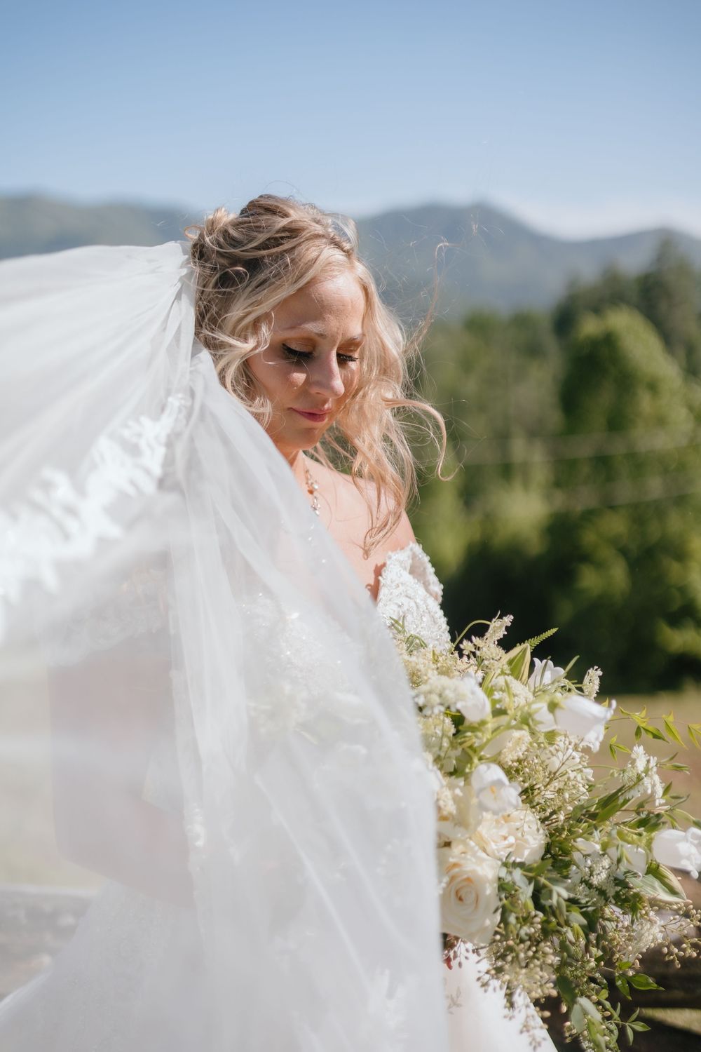 A bride in a flowing white veil poses outdoors with a lush mountain landscape and delicate white floral bouquet in view.