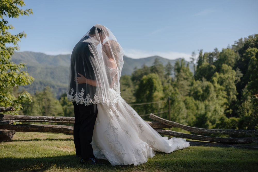 A bride in a flowing white veil and gown stands against a scenic mountain backdrop on a sunny day.