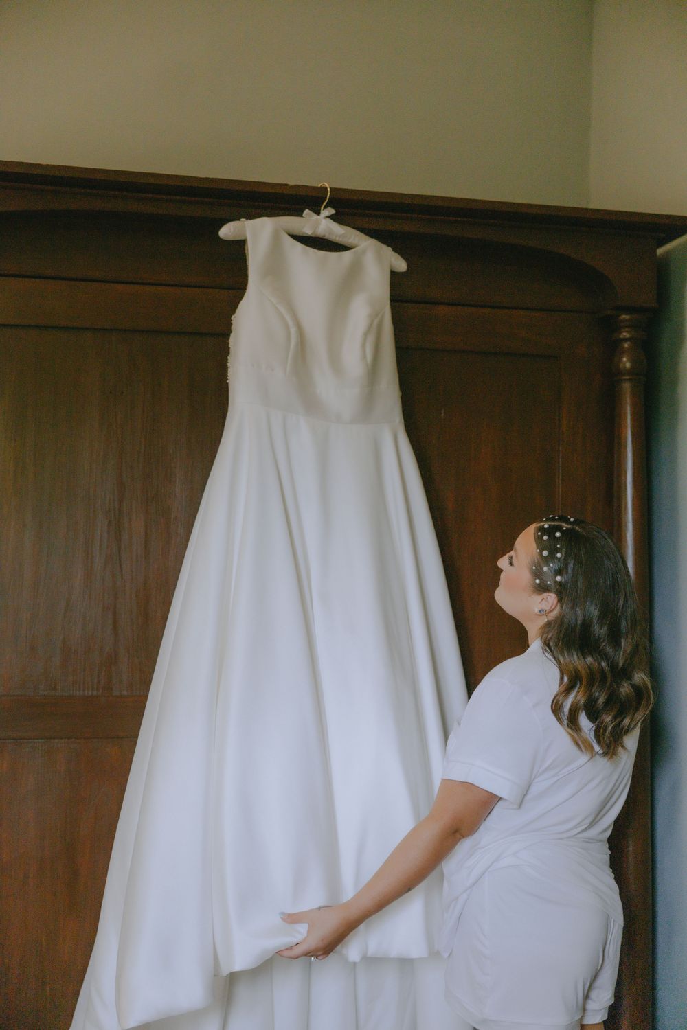 A wedding dress hangs elegantly against dark wooden paneling as someone admires it.