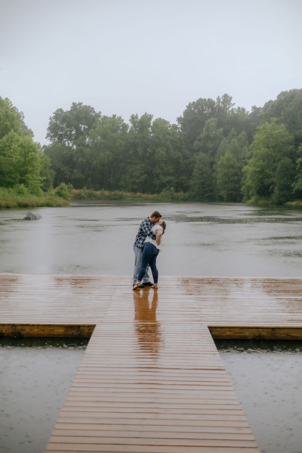 A couple embraces on a wooden dock over a misty lake surrounded by green trees on a cloudy day.