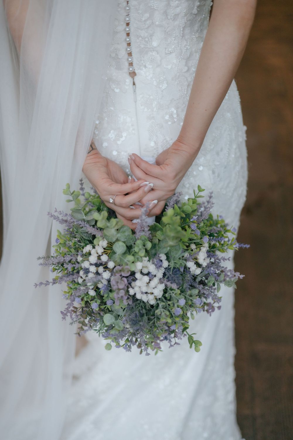 A wedding bouquet with delicate purple and white flowers mixed with green foliage against a white dress background.