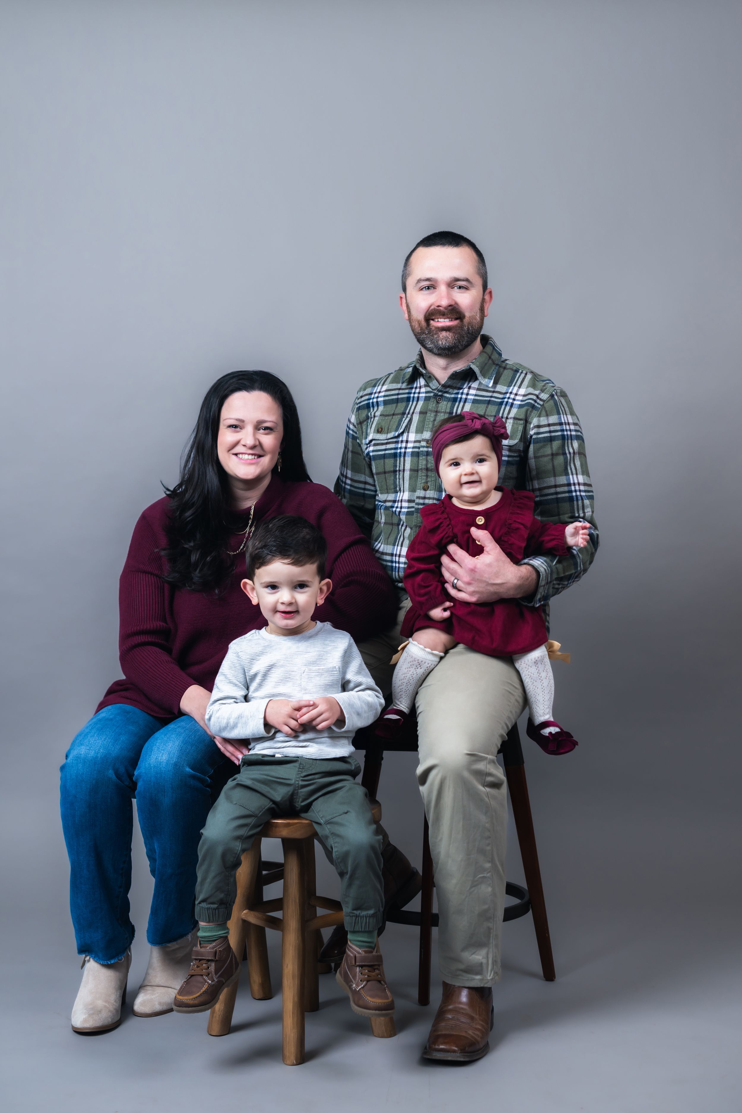 Family of four posing for a studio portrait, parents seated with young son and baby daughter in maroon outfits.