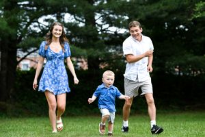 A family runs and plays together on a grassy lawn with pine trees in the background.