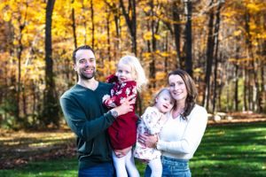 A family group stands together outdoors against a backdrop of fall foliage.
