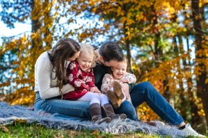 A family shares a playful moment while sitting on grass with autumn trees in the background.