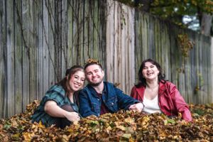 Three friends sitting together in fallen leaves against wooden fence backdrop.