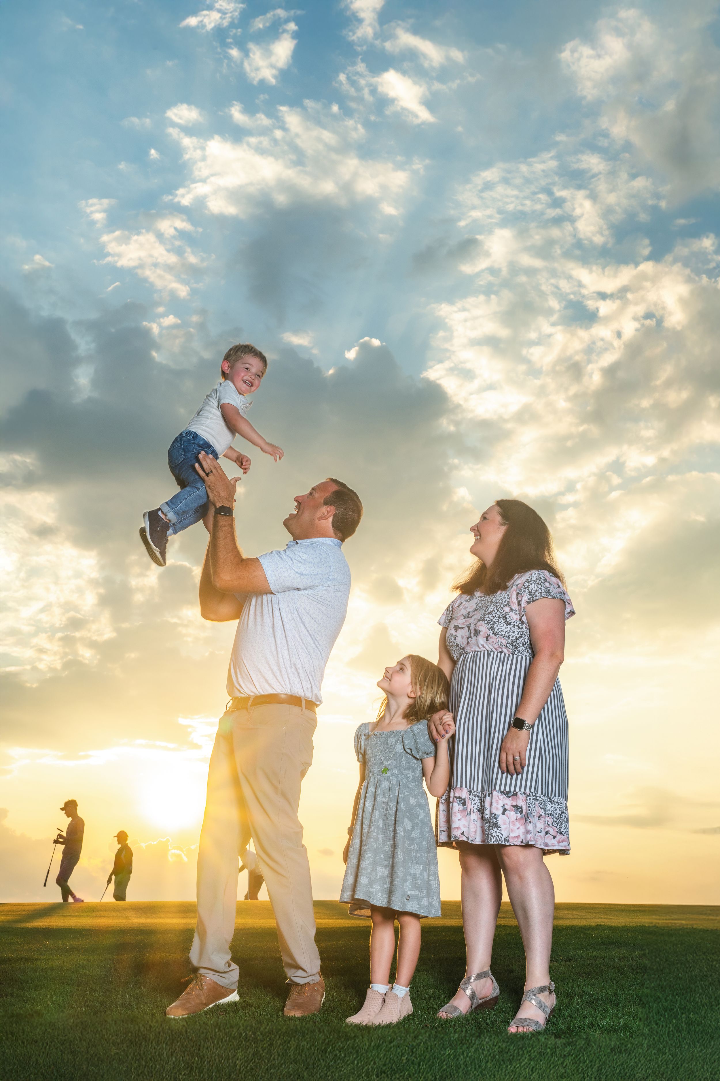 Family enjoying sunset outdoors, father lifting toddler overhead while mother and daughter watch joyfully on green lawn.