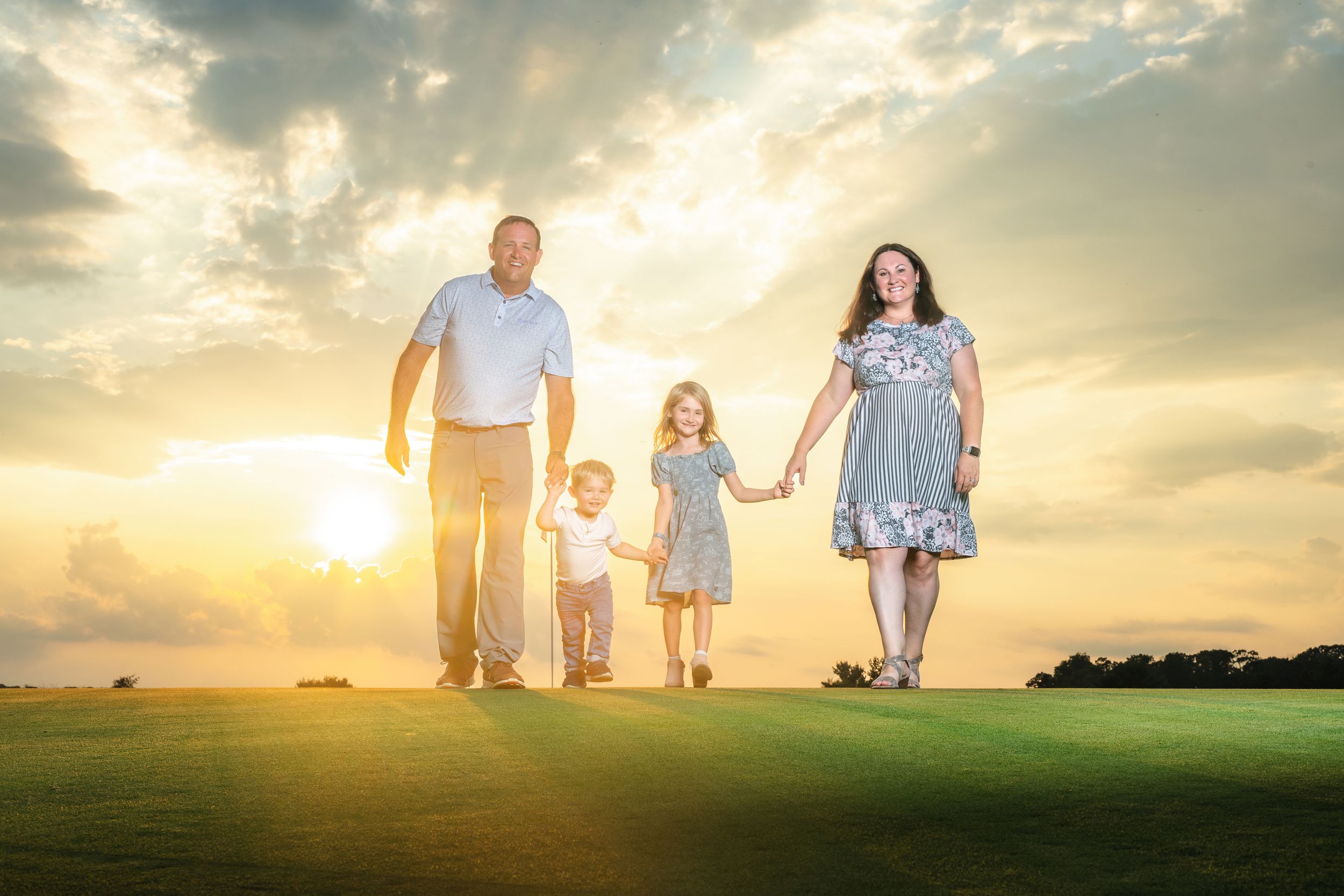 Family of four walking on green grass at sunset with dramatic sky in background.