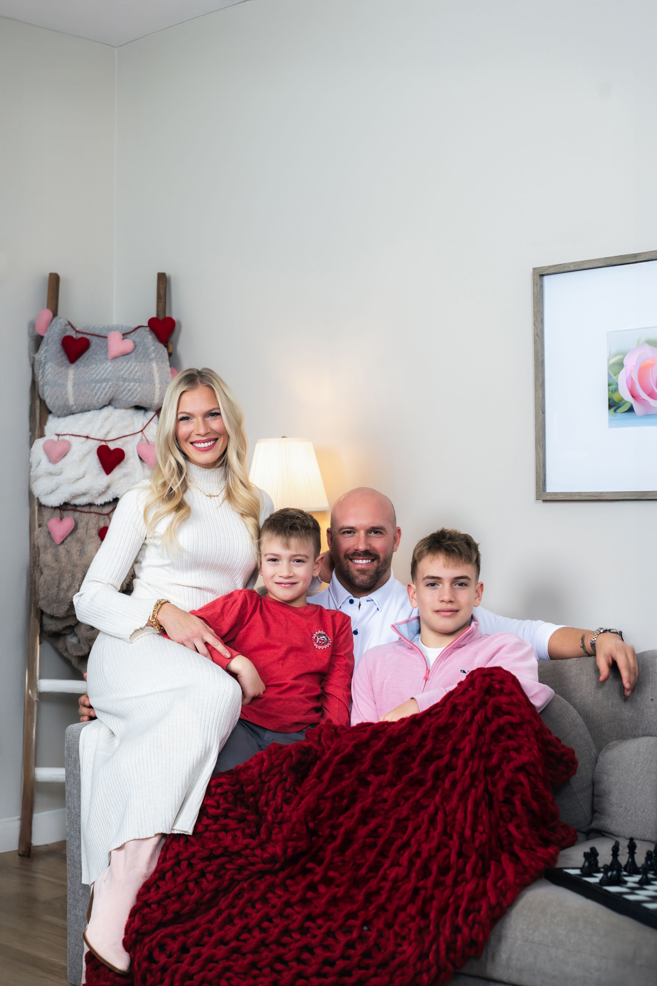 Happy family of four posing together on a couch with a red chunky knit blanket and Valentine's Day decor.