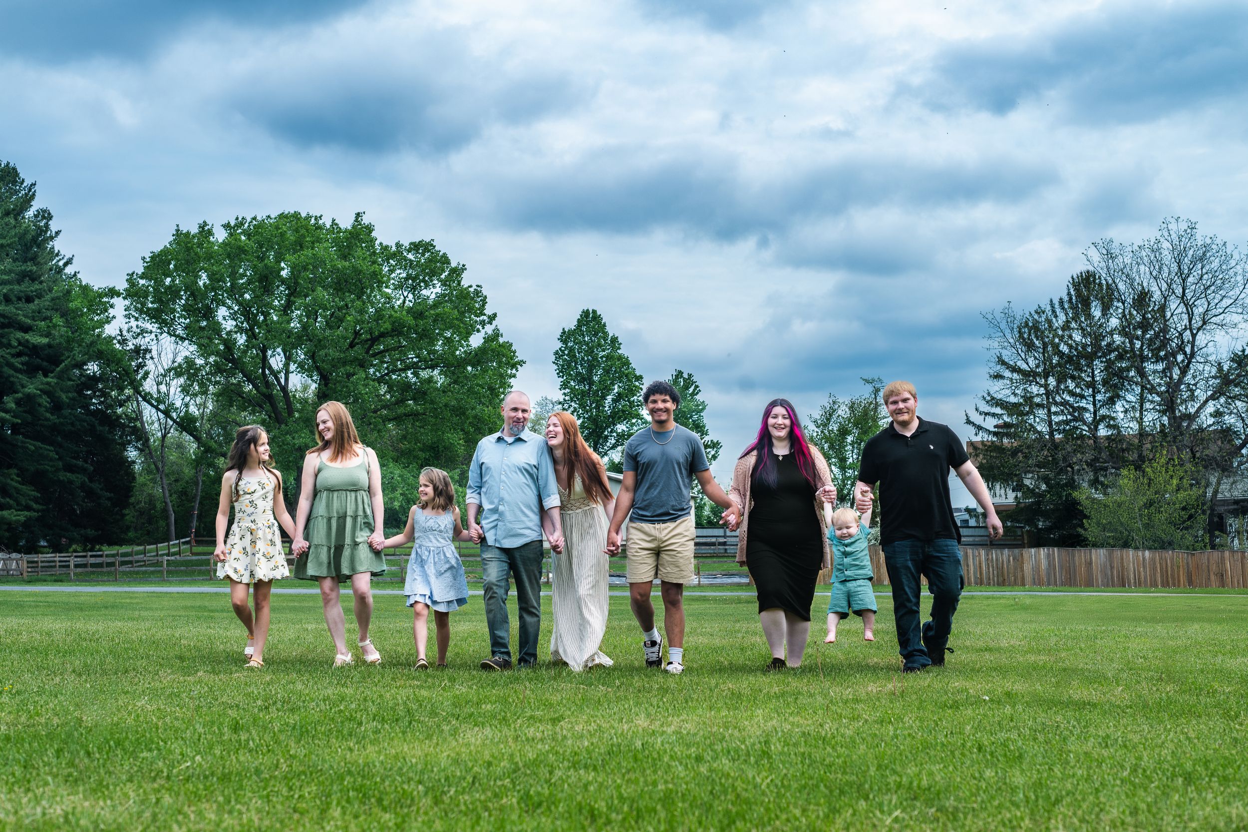 Large family holding hands walking across green grass field under cloudy sky outdoors.