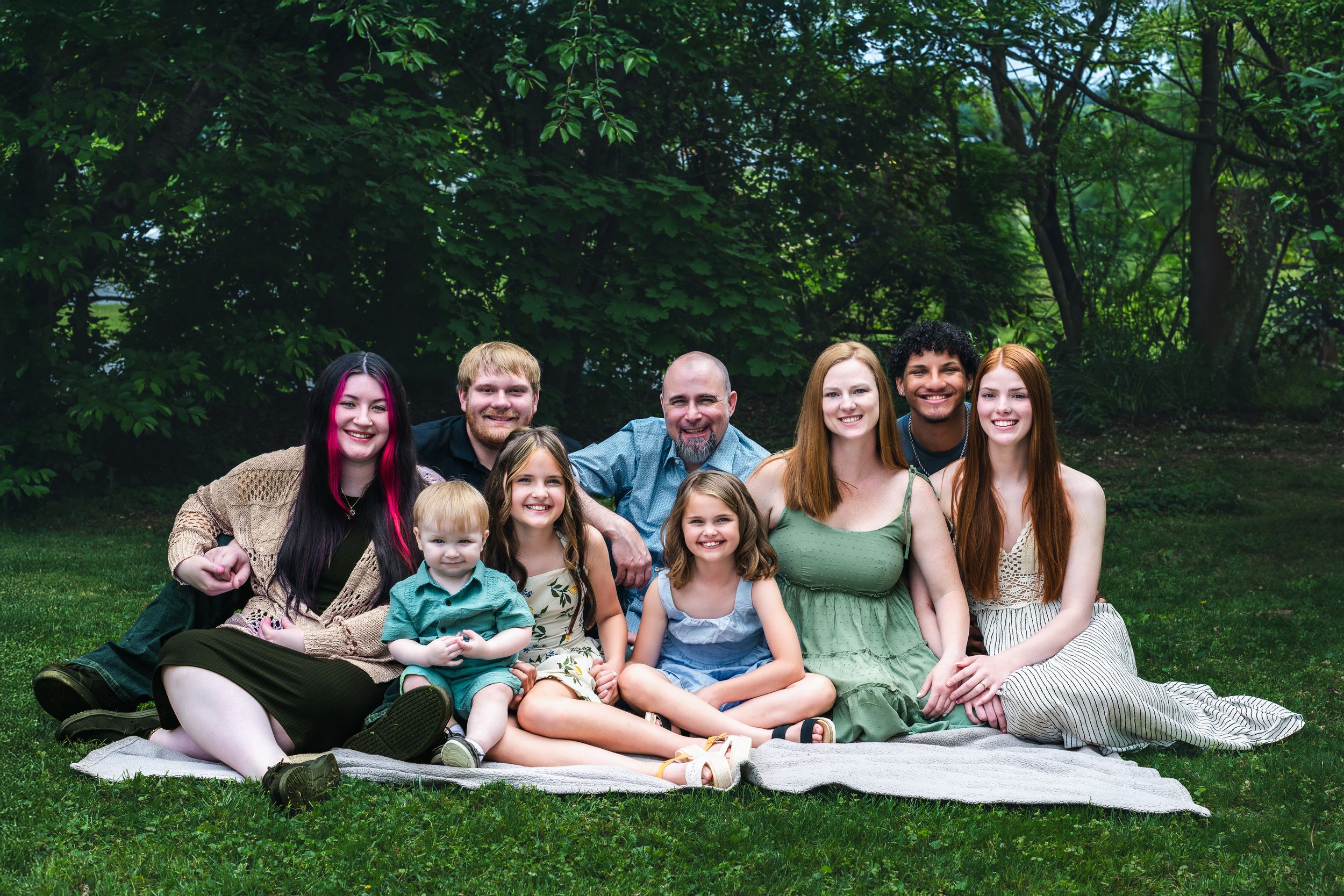 Large family group portrait smiling outdoors on grass in a lush green park setting on a sunny day.