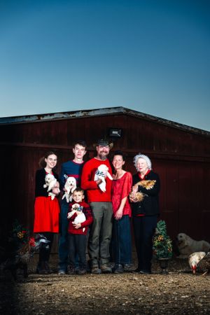 Family wearing red and white poses with puppies outside rustic barn structure.