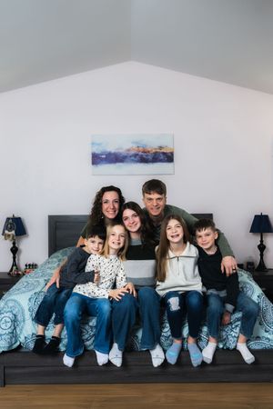 Happy family of seven posing together on a bed, smiling at the camera in a bright bedroom.