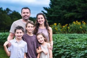 Family of five poses together in front of lush garden with sunflowers in background.