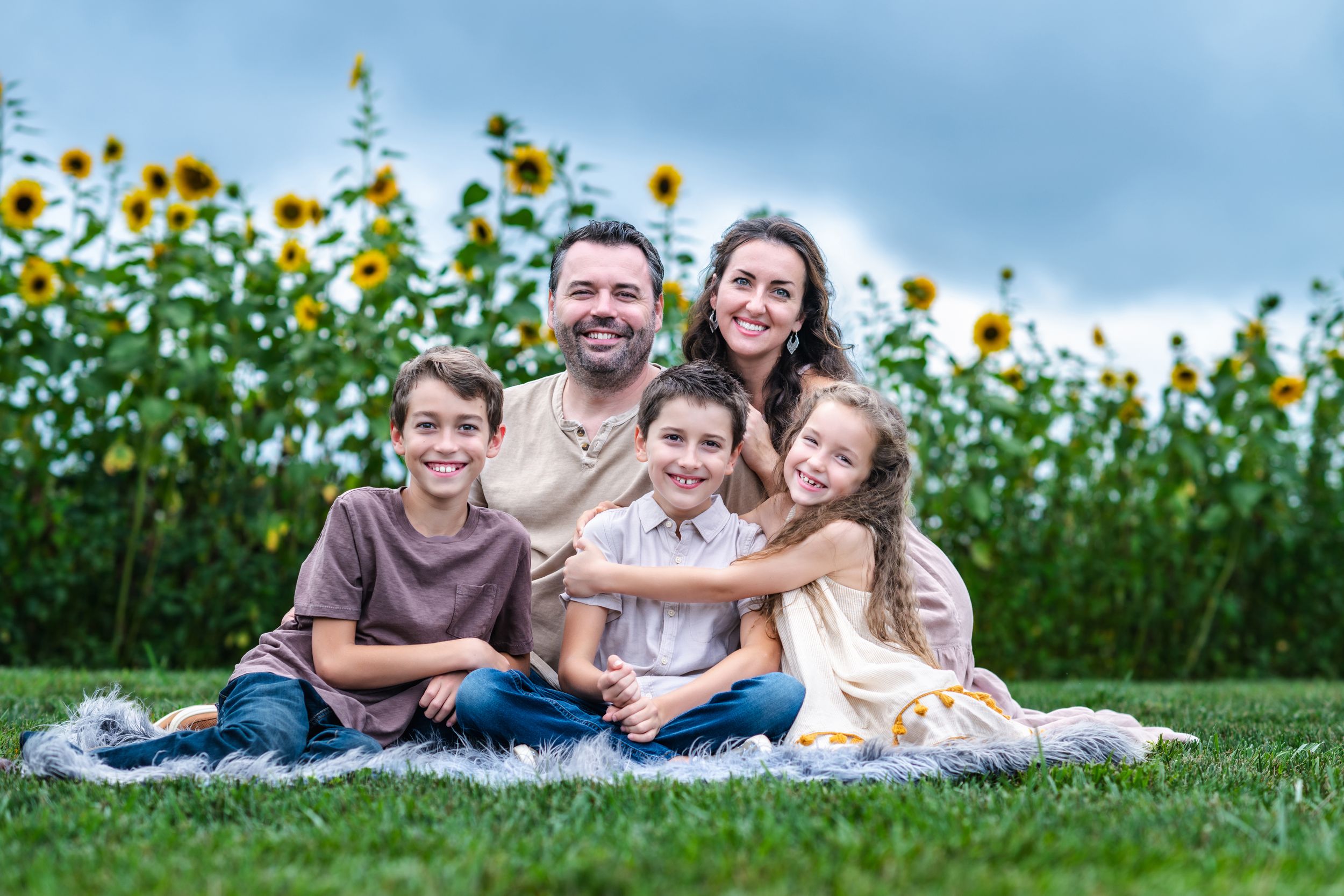 Family sitting on blanket in front of tall sunflower field during golden hour.