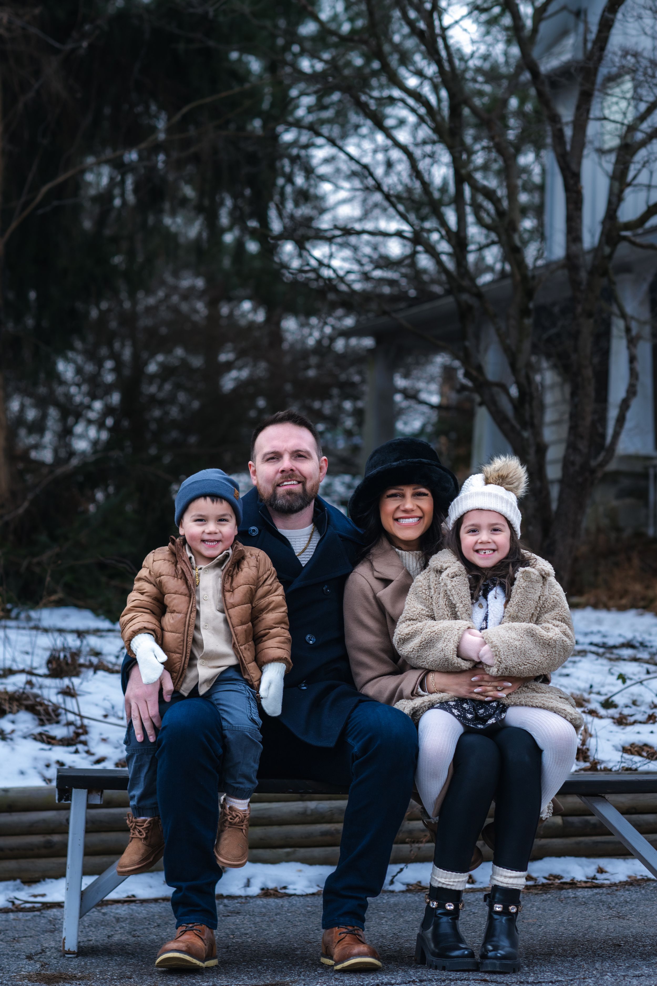 Family of four smiling on a bench in a snowy park, wearing winter coats and hats on a cold day.