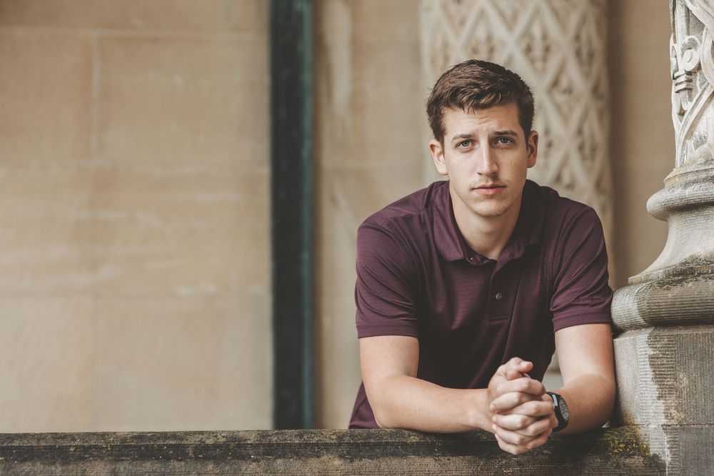 Young man in burgundy polo shirt leans on stone railing in front of decorative architectural background.