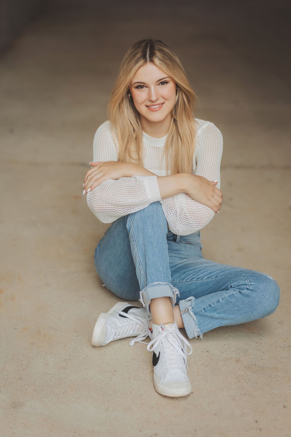 A person in a white knit sweater and blue jeans sits cross-legged in a series of casual poses on a beige floor.