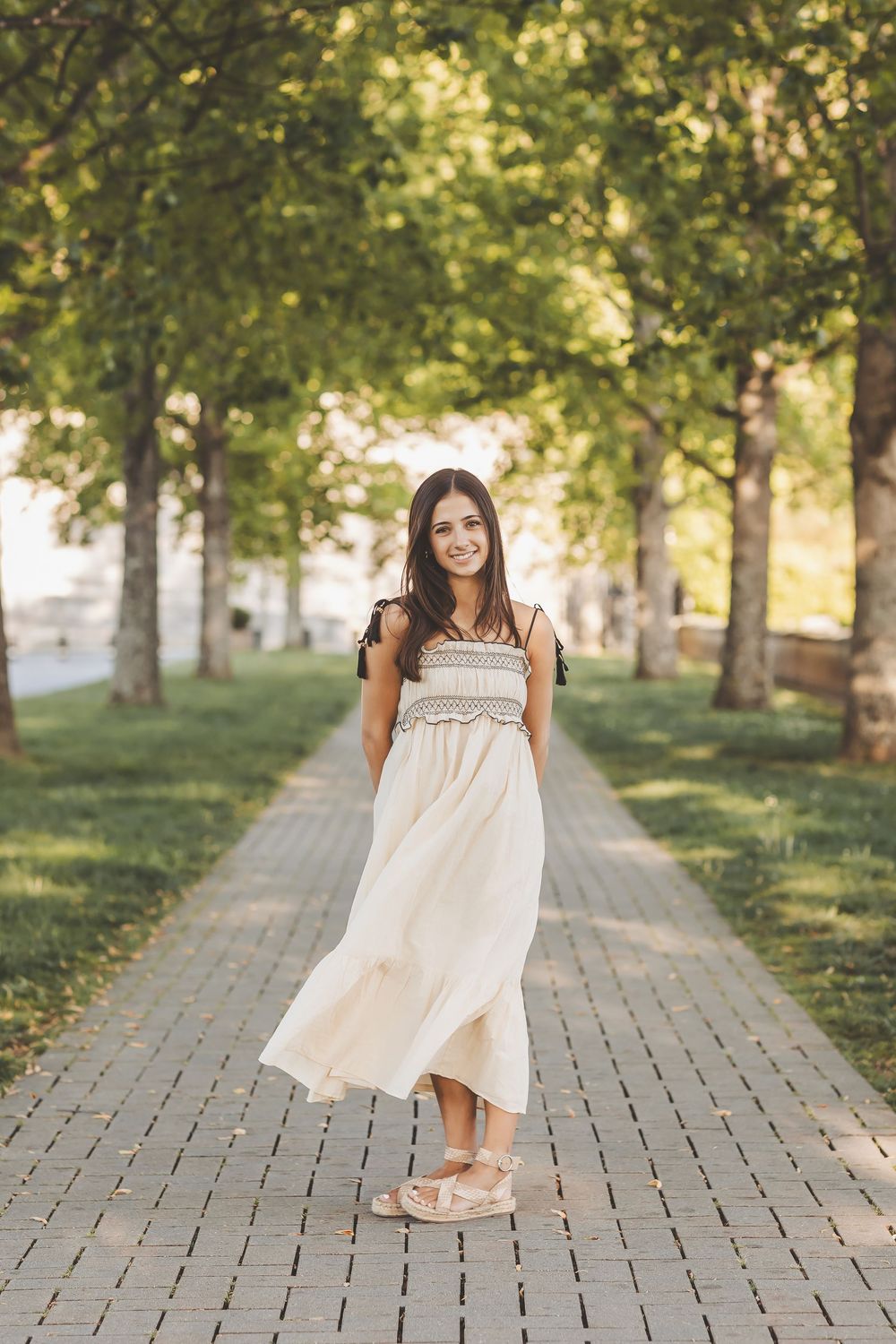 A person in a flowing white summer dress twirls playfully on a brick path surrounded by green trees in golden sunlight.