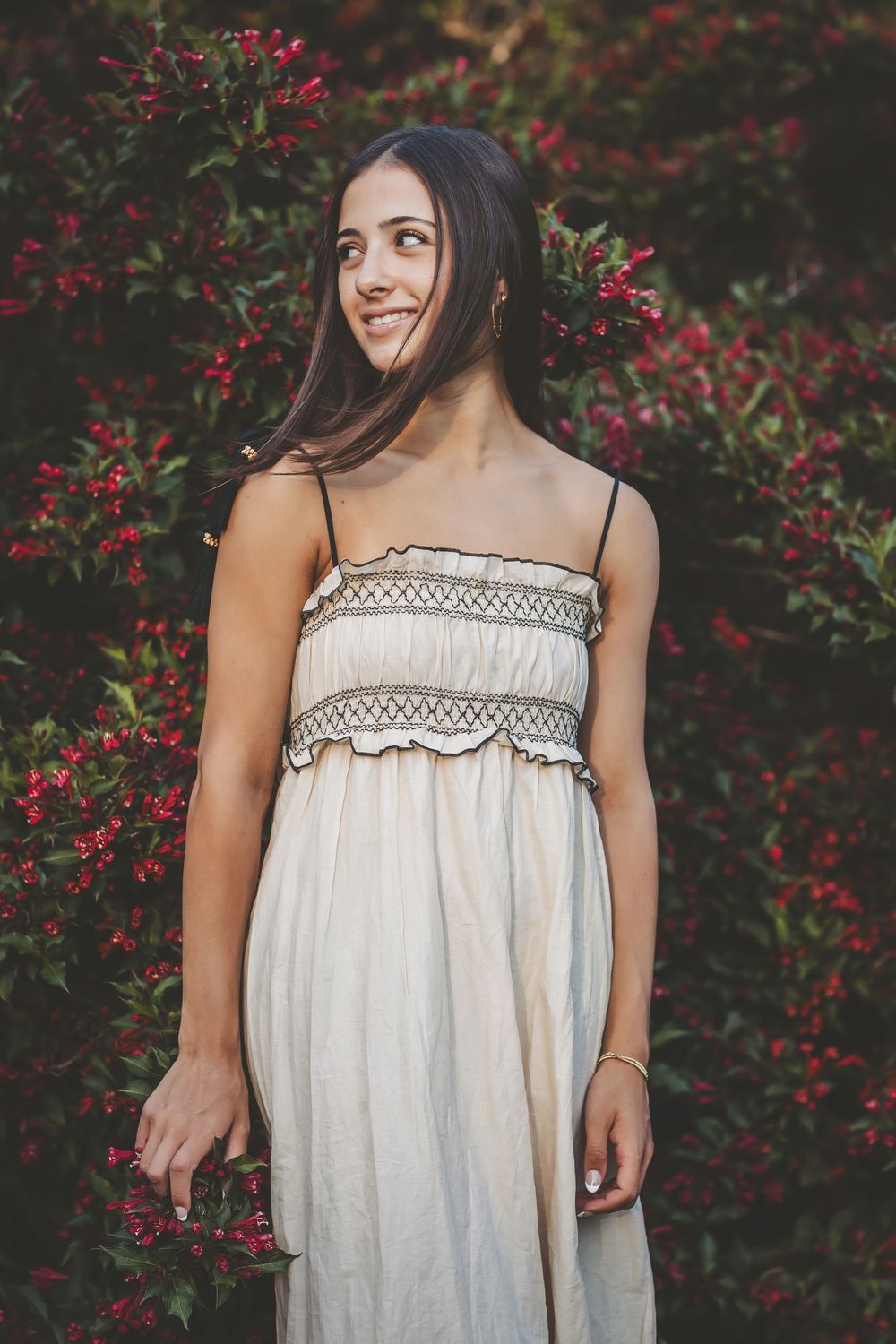 Person in a white summer dress poses against a background of vibrant red berries and lush green foliage.
