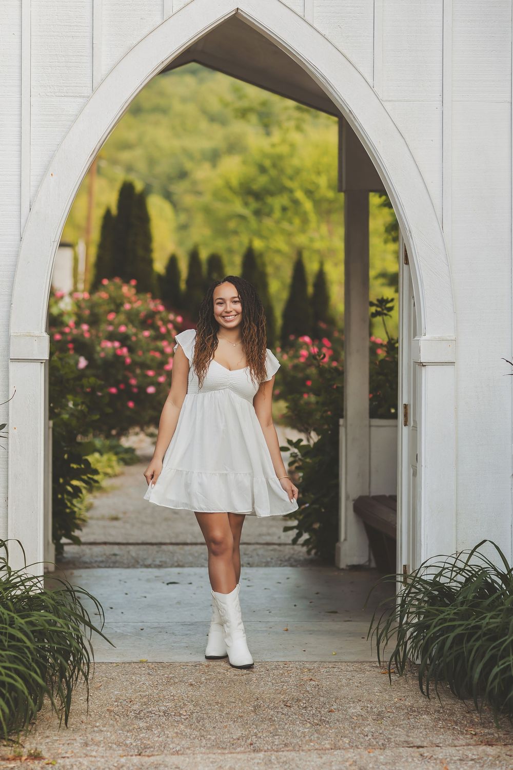 Figure in white dress walking through arched garden pathway with blooming flowers.