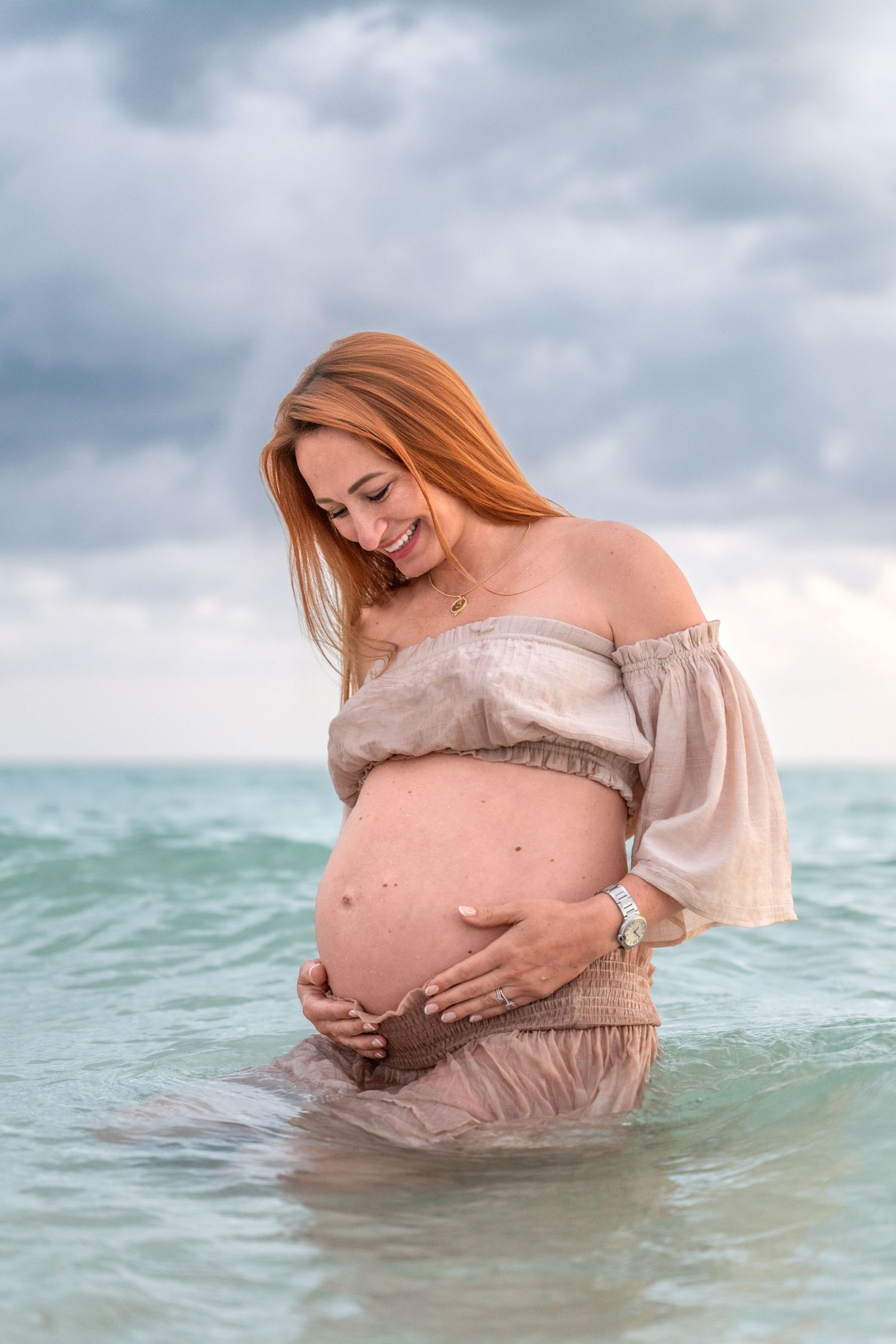 Smiling pregnant woman in off-shoulder dress standing in ocean water, cradling her baby bump on a cloudy day.