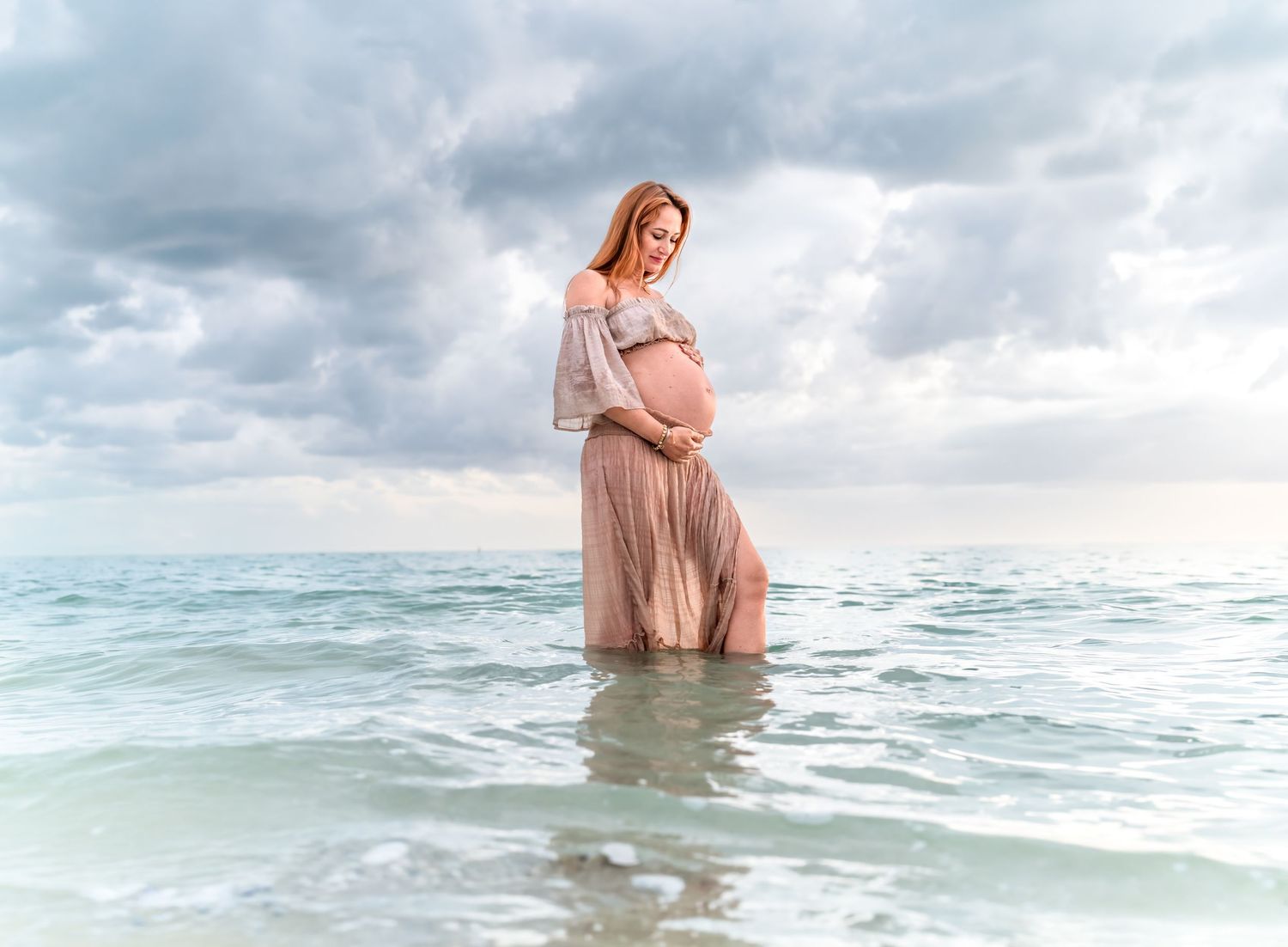 Pregnant woman in pink dress standing in ocean waves under cloudy sky during maternity photo shoot.