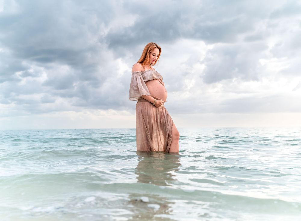 Pregnant woman in pink dress standing in ocean waves under cloudy sky during maternity photo shoot.